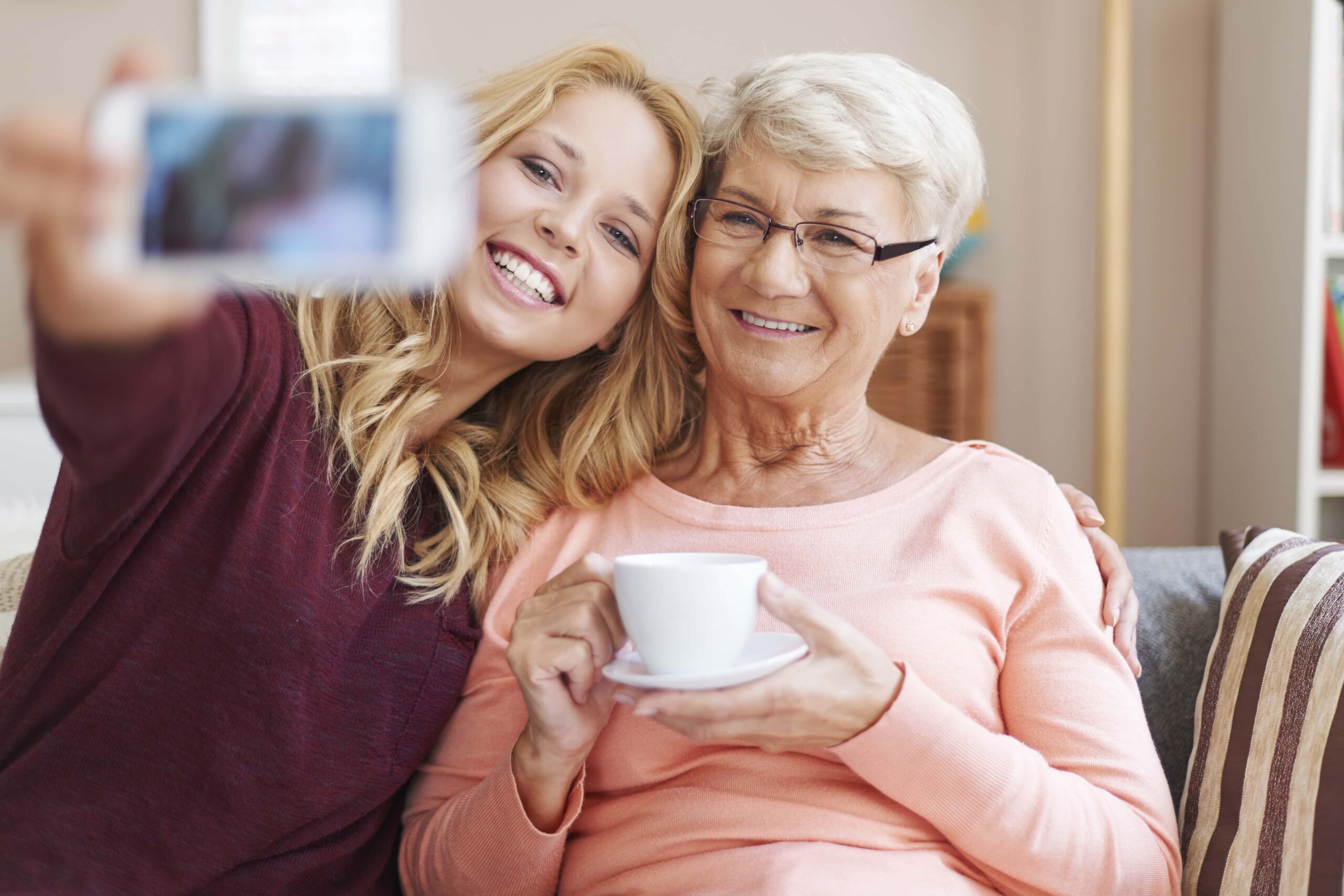 Smiling elderly holding a teacup and smiling younger lady sitting down taking a selfie.