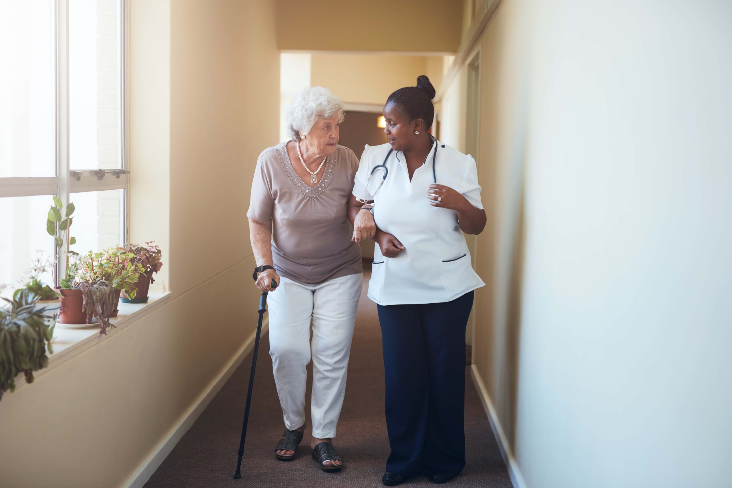 Elderly lady with cane being assisted to walk down an hallway with younger nurse.